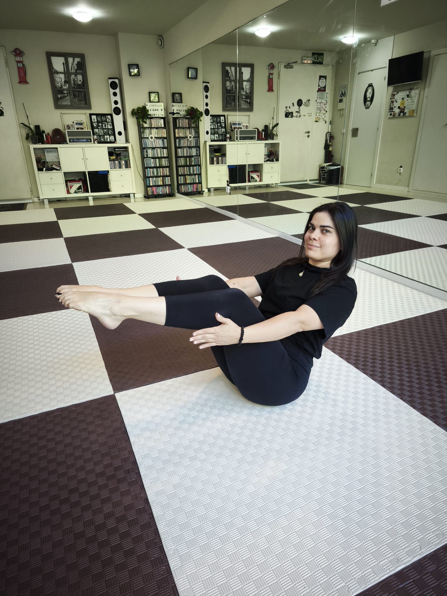 Una alumna en equilibrio sobre los glúteos, con las piernas elevadas y las manos bajo las rodillas, en el suelo ajedrezado.